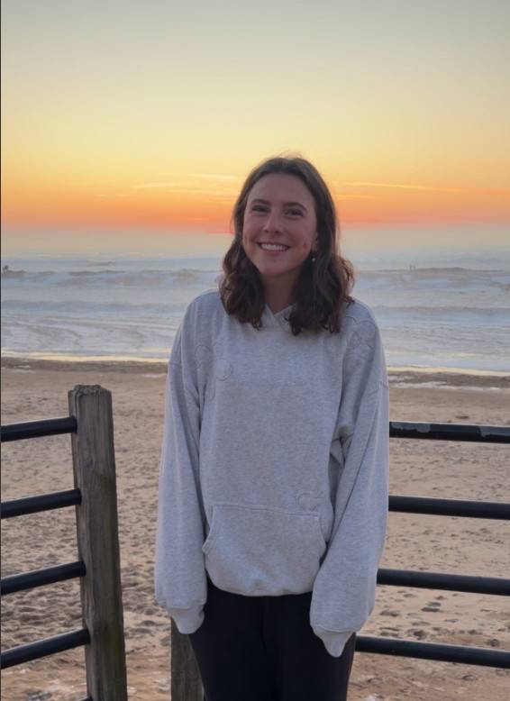 Young woman with brown hair smiling at the camera wearing a grey sweatshirt with a sunset in the background